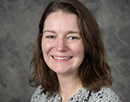 Professional headshot of a person with shoulder-length hair wearing a patterned light-colored blouse. The portrait is cropped from the shoulders up, with the person facing the camera against a mottled gray studio background. The image has a formal academic or professional style