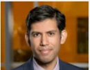 Professional headshot of professor Lav Varshney wearing a dark suit jacket, white dress shirt, and blue tie against a softly blurred indoor background with warm tones.