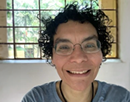 Close-up portrait of professor Liliana Davalos wearing glasses and a light blue shirt, photographed indoors in front of a large window showing a bright outdoor scene with trees visible through the window