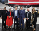 A group of eight adults stands together in an industrial or technology-focused facility, posing for a photo beneath large red, white, and blue equipment or structural elements. Several people wear business attire, while others wear company-branded work clothing