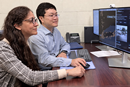 Two researchers sit at a desk collaborating in front of dual computer monitors. One researcher uses a mouse while both look at data visualizations and software displayed on the screens. A keyboard, office phone, and documents are visible on the desk in an office setting