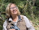 Distinguished service professor Patricia Wright wearing glasses and an outdoor field vest holds binoculars while standing in a wooded natural environment. Trees and dense vegetation fill the background, emphasizing a wildlife research or field observation setting