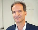 Professional headshot of a person wearing a navy blazer and light-colored shirt, photographed indoors against a softly blurred background with a whiteboard visible behind them