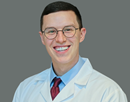 Professional studio headshot of resident Seth Weir wearing glasses, a white medical coat, a light blue dress shirt, and a red-and-blue striped tie against a plain gray background