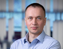 Professional headshot of associate professor Stanley Bak wearing a light blue button-down with the background softly blurred, showing vertical window blinds and an office-like indoor setting