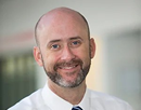 Professional headshot of professor Timothy Glotch from Stony Brook University with a closely trimmed beard, wearing a white dress shirt and blue striped tie against a softly blurred indoor background