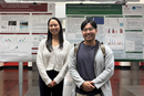 Two researchers stand side by side in front of large scientific posters displayed at a research presentation event. The posters contain charts, graphs, photographs, and text organized into multiple sections. The researchers are positioned in the foreground of a bright indoor atrium with red flooring and additional poster displays visible in the background