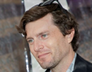 Close-up portrait of associate professor Daniel Holstein from Stony Brook University viewed from a slight side angle. Sunglasses rest on top of the head, and the person wears a dark jacket over a light collared shirt. The background is softly blurred, drawing attention to the face