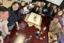 A group of students and faculty gather around a large table during a hands-on study session. At the center of the table is a framed botanical specimen, surrounded by preservation materials, sample containers, and research tools. The participants are positioned around the display, looking toward the camera in a classroom or laboratory setting