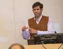 A person stands at the front of a classroom or lecture hall, speaking beside a computer monitor and podium. The person wears a light button-down shirt with a brown knitted sweater vest and gestures with both hands while presenting. A whiteboard fills the background, and part of the audience is visible in the foreground