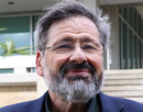 Person with gray hair, beard, and glasses wearing a dark blazer, standing outdoors in front of a building