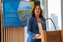 Speaker stands at a podium delivering remarks during an event, with a screen behind displaying information about the New York Climate Exchange. Large windows and a city skyline are visible in the background