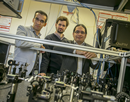 Three people stand behind a complex laboratory apparatus filled with optical components, metal mounts, and precision instruments. The equipment occupies the foreground, emphasizing a research setting focused on advanced physics or engineering