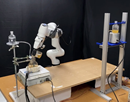 Laboratory robotics setup featuring a robotic arm mounted on a workbench between two metal support frames. The arm is positioned above a platform containing equipment and sensors, with cables and electronic components connected throughout the system. The apparatus is arranged in a controlled indoor research environment with a dark backdrop, highlighting robotics and automation technology