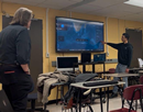 Student presents satellite or mapping data on a large classroom display while an instructor observes. Computers, desks, and classroom equipment are visible throughout the room