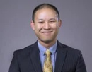 Professional headshot of a person wearing a dark suit and patterned gold tie against a gray studio background