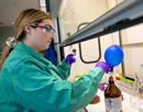 A young woman wearing a green lab coat and purple gloves dips an instrument with a long wire probe into a brown bottle with a small blue balloon attached to its mouth