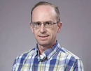 Professional headshot professor Paul Roundy from University at Albany wearing glasses and a blue-and-white plaid button-down shirt against a plain light gray background