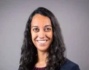 Professional headshot of a person with long dark hair wearing a navy blazer and purple top against a gray studio background