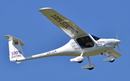 A small white electric airplane flies against a clear blue sky. Bold text reading “100% Electric” is visible on the wing and tail, and additional markings on the fuselage identify the aircraft as an experimental electric-powered plane. The image highlights advances in sustainable aviation technology and zero-emission flight