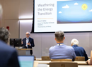 Presenter speaks to an audience in a lecture hall while a presentation slide displaying an energy-related topic is projected on a large screen. Attendees are seated facing the speaker