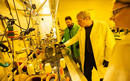 Three researchers wearing lab coats, safety glasses, and protective gloves work around specialized equipment in a laboratory. One researcher points toward an instrument on a crowded laboratory bench while two others observe. Tubing, glassware, gauges, and containers are visible throughout the workspace, which is illuminated by bright yellow lighting commonly used in nanotechnology or semiconductor