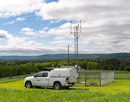 Weather monitoring station and instrument tower in a grassy field, with a research vehicle parked nearby and rolling hills visible in the background
