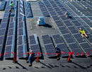 Aerial view of workers installing rows of solar panels on a large rooftop. Several workers wearing high-visibility safety vests are positioned throughout the array, assembling and securing the panels. The dark blue solar panels form a geometric grid pattern that covers much of the roof, highlighting a renewable energy installation project