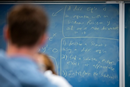 Person viewed from behind while looking at a chalkboard covered with mathematical equations and formulas. The chalkboard fills most of the image, with handwritten calculations in white chalk on a blue surface, suggesting a classroom, teaching, or mathematics-learning environment