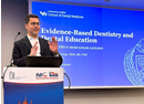 Presenter speaking at a podium beside a slide titled “Evidence-Based Dentistry and Dental Education” at a conference