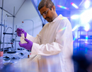 A researcher wearing a white lab coat, safety glasses, and purple gloves works at a laboratory bench, carefully handling a flask and laboratory tool. Glassware, instruments, and scientific equipment are arranged across the workspace, while bright blue and rainbow-colored reflections from laboratory lighting create a vivid backdrop