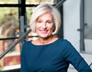 Professional headshot of a person with short, light-colored hair wearing a long-sleeved blue top positioned indoors near large windows and a staircase, with natural light illuminating the scene and has a softly blurred background featuring architectural details