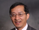 Professional headshot of distinguished professor Chunming Qiao from University at Buffalo wearing glasses, a gray suit jacket, white dress shirt, and red patterned tie set against a neutral gray studio background