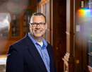 Person wearing glasses and a blue shirt poses in a wood-paneled office or library, with bookshelves and decorative window light visible in the background