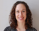Professional headshot of a person with shoulder-length curly hair, wearing a dark blouse with a small white pattern against a plain light-colored background