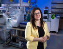 Associate professor Emanuela Giofriddo wearing glasses and a light yellow blazer stands in a laboratory beside the laboratory equipment. Scientific equipment, tubing, and stainless-steel machinery fill the background, while blue-toned lighting highlights the technical workspace