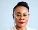 Professional headshot of a person wearing glasses, earrings, and a white blazer against a light blue background