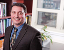 Gregory Homish in business attire poses in an office with bookshelves, a window, and a potted plant visible in the background
