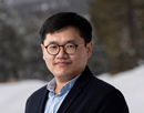 Professional headshot of a person wearing glasses, a dark blazer, and a light-colored collared shirt taken outdoors. The background is softly blurred, showing snow-covered ground and dark trees, creating a winter setting