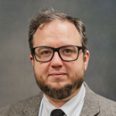 Professional headshot of a person wearing rectangular black eyeglasses, a gray patterned blazer, a light striped dress shirt, and a dark tie against a neutral gray background