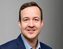 Professional headshot of a professor Jonathan Lovel wearing a blue suit jacket and light blue dress shirt against a gray background