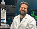 Postdoctural scholar Joseph Basalla in a laboratory coat next to a microscope, with a monitor displaying a fluorescent microscopy image