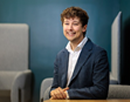 Professional portrait of assistant professor Joshua Miller seated in a modern office setting, wearing a dark blue suit jacket and a light-colored dress shirt against a blue wall, with upholstered seating and privacy panels visible in the background