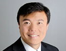 Professional headshot of professor Jun Qu from University at Buffalo wearing a dark suit jacket, white dress shirt, and red patterned tie against a plain light gray background