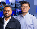 From left, Harim Tavares dos Santos and Zhibin Cui standing in a laboratory with shelves of scientific equipment and sample containers in the background. The image highlights a collaborative research setting with lab instruments illuminated by blue lighting