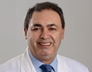 A smiling man with short dark hair wearing a white lab coat over a light blue shirt and patterned blue tie, photographed against a plain light gray background