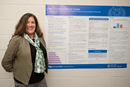 A person stands beside a large academic research poster mounted on a display board. The poster contains multiple sections of text, charts, and graphs arranged in columns. The person is wearing a blazer over a patterned blouse and is positioned next to the presentation in an indoor conference or research showcase setting