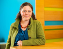 Professor Sophie Nowicki from University at Buffalo wearing a green jacket over a blue top and a beaded necklace stands beside a wooden table in a brightly lit office or campus setting with a turquoise wall and decorative wood paneling with colorful horizontal accents background