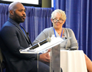 Two presenters stand at a lectern during a conference session. One presenter speaks into a microphone at the podium while the other stands beside a high-top table. A projection screen and blue stage curtains are visible in the background, indicating a professional academic or scientific conference setting