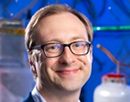Professional headshot of a person wearing glasses and a blue jacket, photographed in a laboratory setting with scientific equipment in the background